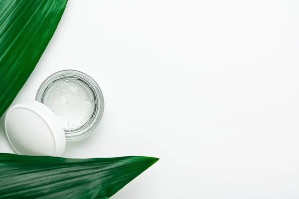 A transparent face care gel in a small jar on large green tropical leaves. Cosmetic beauty product for moisturizing face, hands and body skin copy space. Natural organic cosmetic. Top view, flat lay.