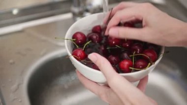 Böğürtlen işleme. Kadın elleri, beyaz kaseyi kırmızı kirazlı tutarak akarsuyun altında yıkanır ve evdeki yakın görüşte mutfaktaki gri lavaboya su döker.