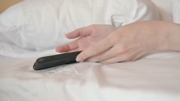 女性的手伸出手去拿现代智能手机 而女性则躺在舒适的床上 在网上冲浪 上网成瘾问题 — 图库视频影像