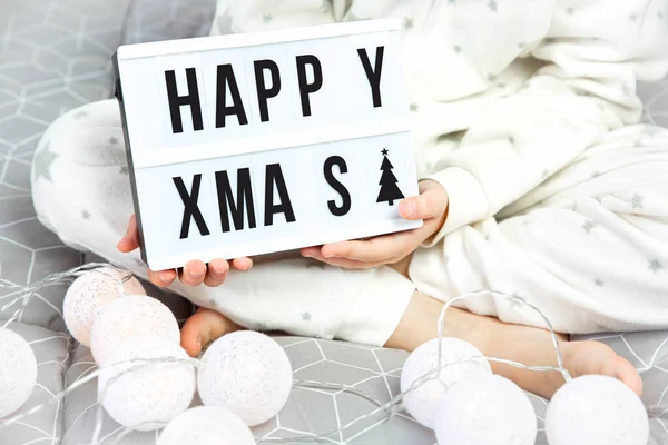 Un niño en pijama blanco se sienta con las piernas cruzadas en la cama y sostiene una tabla ligera con la inscripción feliz Navidad en sus manos. — Foto de Stock