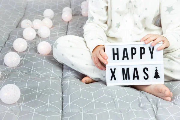 Дитина в білій піжамі сидить хрестоподібно на ліжку і тримає світлу дошку з написом щасливе Різдво в руках . — стокове фото
