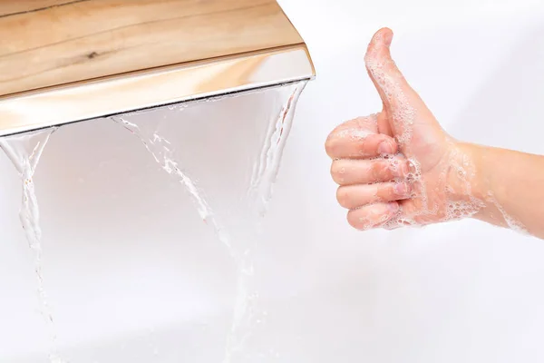 Lávese las manos con jabón bajo un grifo con agua limpia. jabonosa mano de los niños muestra el pulgar de cerca sobre un fondo blanco. — Foto de Stock