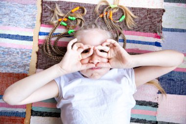 Yakın plan portre örgülü saçları olan neşeli bir kız, örülmüş bir kilimin üzerinde yatıyor, gülümsüyor, katlanmış parmaklarından gözlük takıyor..