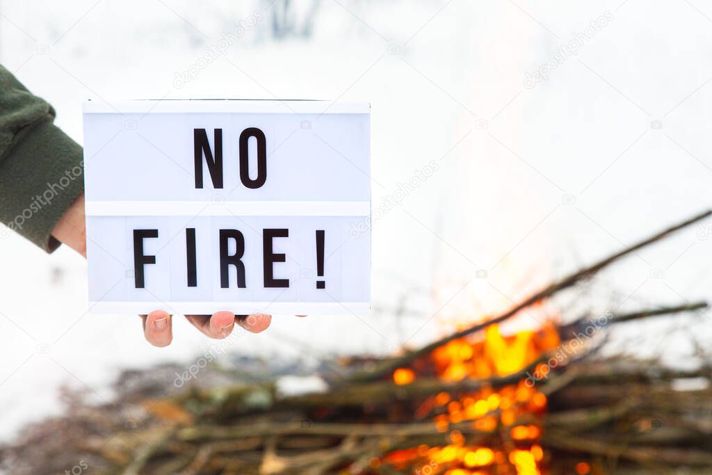 A sign with the text no fire in a mans hand over a flaming bonfire on a white background.