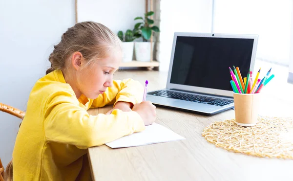 Schoolgirl in a yellow sweatshirt is doing homework at home by the window. — Stock Photo, Image