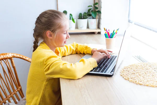 Niña de la escuela caucásica escribiendo en un teclado portátil. Acogedor lugar de trabajo por la ventana. — Foto de Stock