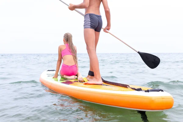 Niño y niña en el tablero de paddle en la tranquila superficie del mar moviéndose hacia el horizonte. — Foto de Stock