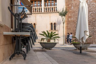 Campos, Balear Adaları / İspanya; Kasım 2020: Campos kentinde kapalı bir kafeterya terası olan ticari cadde. Sandalyeler ve masalar zincirle bağlanmış. Arka planda iki kadın var.