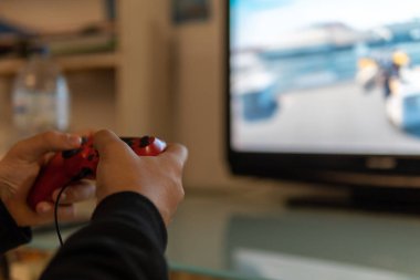 Gençlerin elleri kırmızı uzaktan kumandalı bir konsolla oynuyor ve televizyonun arka planı bulanık.