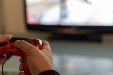 Gençlerin elleri kırmızı uzaktan kumandalı bir konsolla oynuyor ve televizyonun arka planı bulanık.