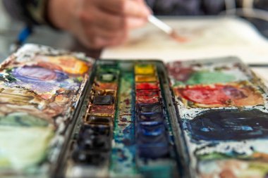 Close-up of a watercolor palette with different colors with a hand drawing in the background out of focus