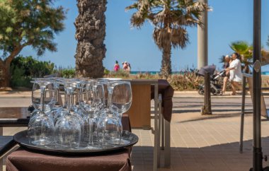 Cam şarap kadehleri, Palma de Mallorca 'daki sahil gezinti alanındaki bir restoranın açık masasına geometrik olarak monte edilmiş ve turistler odak noktasından uzaklaşıp arka planda geziniyorlar.