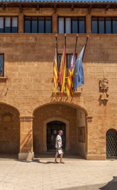 Campos, İspanya; 12 Haziran 2021: Majorcan kasabası Campos belediye binasının ana cephesi, yüz maskesi takan ve Balear Adaları, İspanya ve Avrupa bayrakları altında yürüyen bir turist