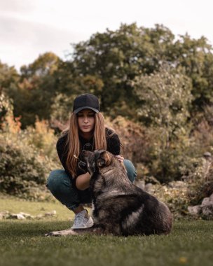 Esmer bir kadının, elinde sopayla bir sahanın ortasında çömelmiş sokak köpeğiyle oynarken dikey çekimi. Kahverengi renkler sıcak bir sonbahar gününde baskın gelir.