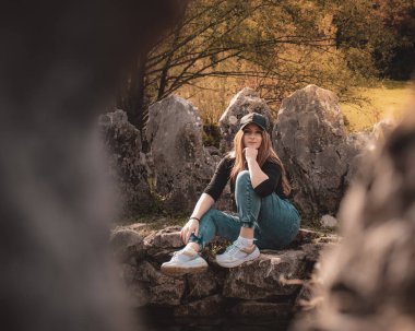 Rajcica Kuyuları 'ndaki bir kuyuda oturan çekici bir esmerin tüm vücut portresi. Kotlu ve siyah şapkalı bir kız, sıcak kahverengi bir sonbahar gününde modern trend kıyafeti