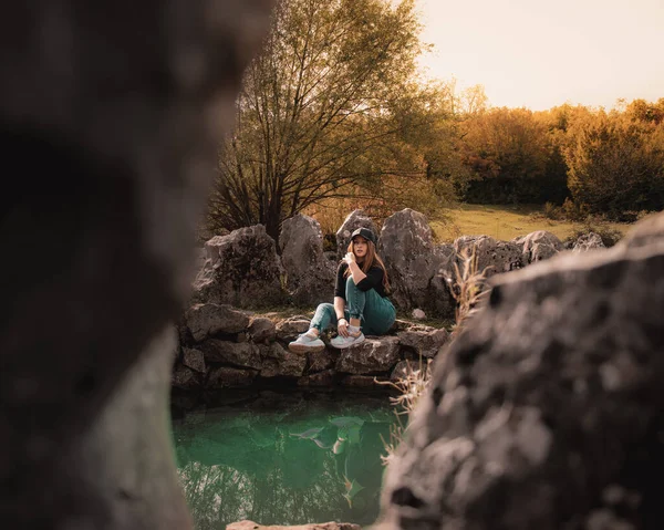 Wide portrait of an attractive brunette sitting on a well at the Rajcica wells natural landmark. Girl wearing jeans and a black cap, modern trending outfit on a warm brown autumn day