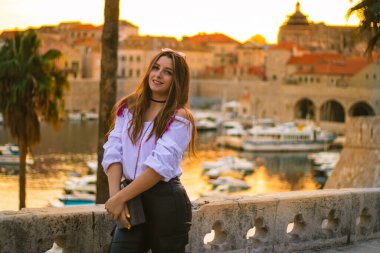 Altın saat boyunca güneş batarken Dubrovnik şehrinin şehir duvarlarında poz veren şık, çekici esmer kadın. Uzaktaki küçük liman güzel turuncu ışıkla aydınlanıyor.