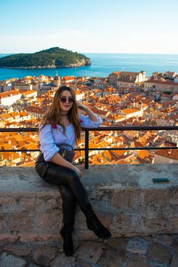 Dubrovnik 'in şehir duvarlarında oturan çekici esmer etkileyici, sosyal medya için dikey bir fotoğraf için poz veriyor. Parlak bir sonbahar gününde arka planda eski şehrin çatıları.
