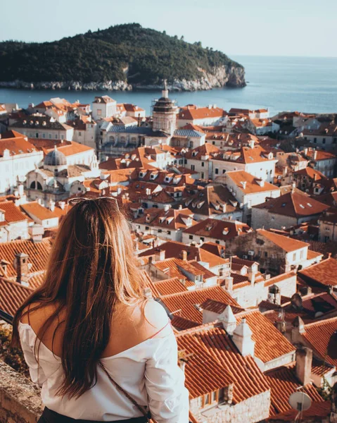 Vertical rear view shot of a girl standing on the city walls of Dubrovnik, looking towards the old red rooftops and the churches in the tightly packed city. Adriatic sea and islands in the distance