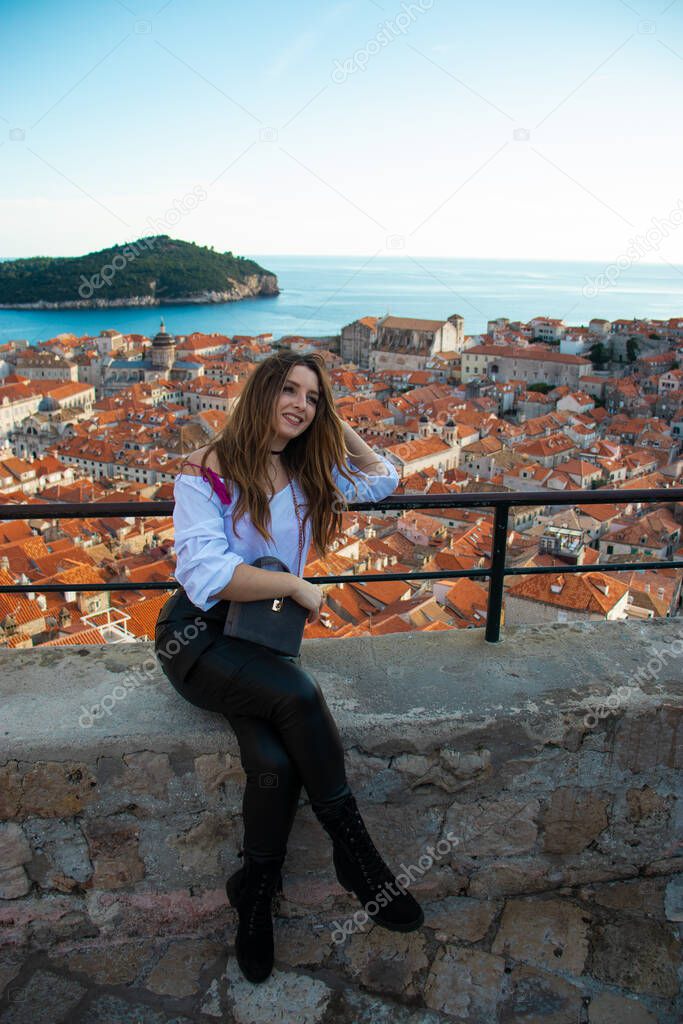 Attractive brunette influencer sitting on the city walls of Dubrovnik, posing for a vertical photo for social media. Rooftops of the old city in the background on a bright autumn day