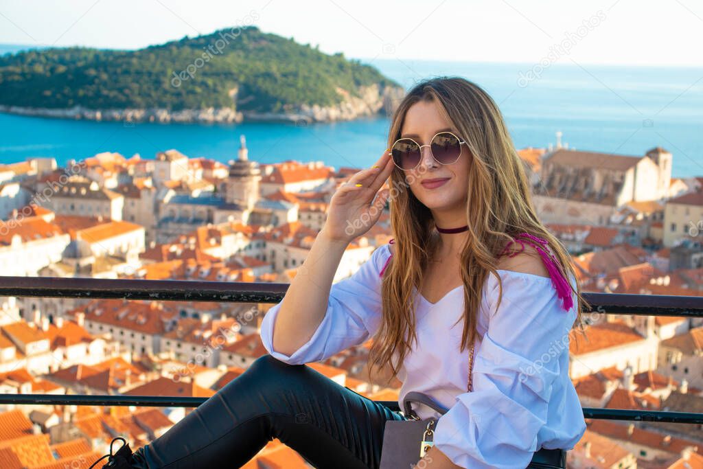 Attractive brunette influencer sitting on the city walls of Dubrovnik, posing for a vertical photo for social media. Rooftops of the old city in the background on a bright autumn day