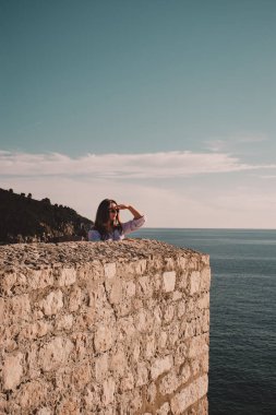 Dubrovnik kasabasının duvarlarında dikilen bir esmerin dikey görüntüsü. Elini gözlerinin üstüne koyup sanki bir şey arıyormuş gibi uzaklığa bakıyor.