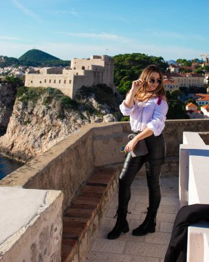Sıcak bir sonbahar gününde antik şehri keşfeden turist Dubrovnik 'in eski şehir duvarlarında poz veren çekici esmer gezgin. Beyaz gömlek ve siyah deri pantolon giyiyor.