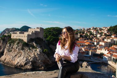 Sıcak bir sonbahar gününde antik şehri keşfeden turist Dubrovnik 'in eski şehir duvarlarında poz veren çekici esmer gezgin. Beyaz gömlek ve siyah deri pantolon giyiyor.