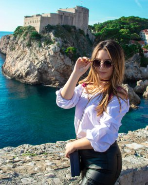Sıcak bir sonbahar gününde antik şehri keşfeden turist Dubrovnik 'in eski şehir duvarlarında poz veren çekici esmer gezgin. Beyaz gömlek ve siyah deri pantolon giyiyor.
