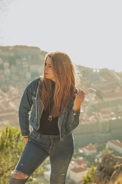 Çekici bir esmerin dikey portresi, kot pantolon ve bluz giyen, dışarıda saçlarıyla oynayan, Dubrovnik şehrinin üzerinde duran. Uzaktaki ünlü antik şehrin ana hatları.