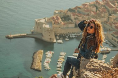 Arkadan görülen çekici esmer, Dubrovnik şehrinin yukarısındaki bir taşın üzerinde poz verirken görülüyor. Arka plandaki görkemli eski kasabanın ana hatları. Duvarları ve Srd dağını ziyaret eden bir gezgin.