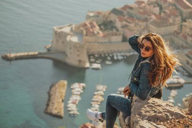 Arkadan görülen çekici esmer, Dubrovnik şehrinin yukarısındaki bir taşın üzerinde poz verirken görülüyor. Arka plandaki görkemli eski kasabanın ana hatları. Duvarları ve Srd dağını ziyaret eden bir gezgin.