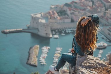 Arkadan görülen çekici esmer, Dubrovnik şehrinin yukarısındaki bir taşın üzerinde poz verirken görülüyor. Arka plandaki görkemli eski kasabanın ana hatları. Duvarları ve Srd dağını ziyaret eden bir gezgin.