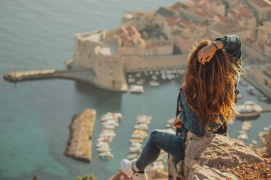 Arkadan görülen çekici esmer, Dubrovnik şehrinin yukarısındaki bir taşın üzerinde poz verirken görülüyor. Arka plandaki görkemli eski kasabanın ana hatları. Duvarları ve Srd dağını ziyaret eden bir gezgin.
