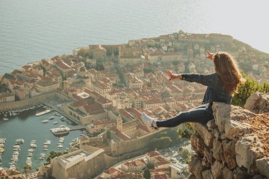 Arkasında Dubrovnik şehri olan bir kayanın kenarında oturan mutlu, heyecanlı kız. Güzel antik şehir öğleden sonra günbatımında parlıyor, Covid mevsiminde ziyaret ediyor.