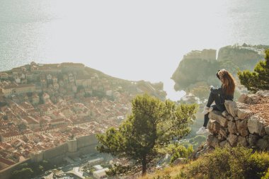 Parlak bir günbatımında bir kayanın kenarında oturan esmer bir kadının geniş açılı görüntüsü. Arkasında eski Dubrovnik kenti var. Dubrovnik 'i ziyaret ederken turist yoktu.