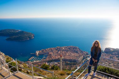 Kot pantolonlu esmer ve Dubrovnik şehrinin yukarısındaki Srd gözlem noktasının korkuluğunda duruyor. Uzaktaki antik şehir mavi Adriyatik Denizi ile çevrili.