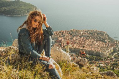 Dubrovnik şehrinin yukarısındaki Srd tepesinde oturan çekici esmer, uzaktan şehri izliyor. Rüzgâr saçlarını antik ve ünlü kasabanın üzerinde savuruyor. Cablecar düşüyor