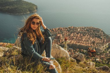 Dubrovnik şehrinin yukarısındaki Srd tepesinde oturan çekici esmer, uzaktan şehri izliyor. Rüzgâr saçlarını esiyor bu tarihi şehrin üzerinde