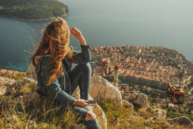 Dubrovnik şehrinin yukarısındaki Srd tepesinde oturan çekici esmer, uzaktan şehri izliyor. Rüzgâr saçlarını esiyor bu tarihi şehrin üzerinde