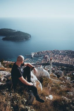 Dubrovnik şehrinin yukarısındaki Srd Dağı 'nda oturan kel bir adamın soğuk bir sonbahar gününde bölgeyi gözlemlediği dikey karamsar bir fotoğraf.
