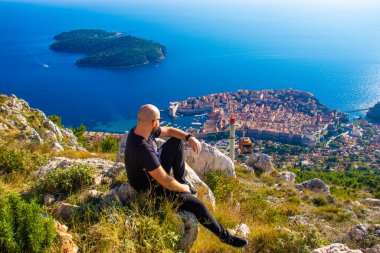 Srd dağının tepesinden geniş açılı çekim, Dubrovnik şehrinin etrafındaki bir kayanın üzerinde oturan adam, turuncu kablocu yazın turist gezginlerini taşıyan şehir duvarlarına iniyor.