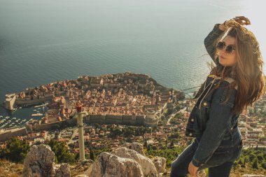 Altın saatinde Dubrovnik şehrinin tepesinde dikilen güzel bir esmerin omuz manzarası. Saçlarını rüzgarda tutan. Dipte, her yıl milyonlarca turist tarafından ziyaret edilen antik ünlü şehir.