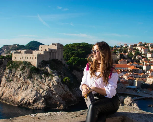 Sıcak bir sonbahar gününde antik şehri keşfeden turist Dubrovnik 'in eski şehir duvarlarında poz veren çekici esmer gezgin. Beyaz gömlek ve siyah deri pantolon giyiyor.