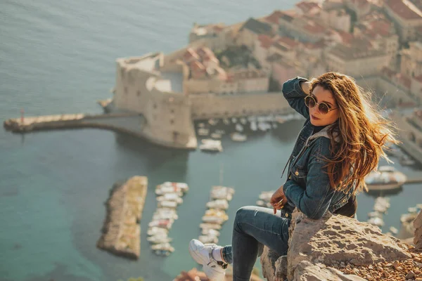 Arkadan görülen çekici esmer, Dubrovnik şehrinin yukarısındaki bir taşın üzerinde poz verirken görülüyor. Arka plandaki görkemli eski kasabanın ana hatları. Duvarları ve Srd dağını ziyaret eden bir gezgin.