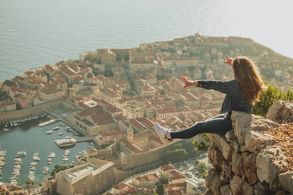 Arkasında Dubrovnik şehri olan bir kayanın kenarında oturan mutlu, heyecanlı kız. Güzel antik şehir öğleden sonra günbatımında parlıyor, Covid mevsiminde ziyaret ediyor.
