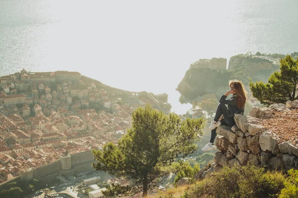 Wide shot of a brunette sitting on the edge of a rock posing during a bright sunset with the old city of Dubrovnik behind her in the distance. Visiting Dubrovnik with no tourists during covid season