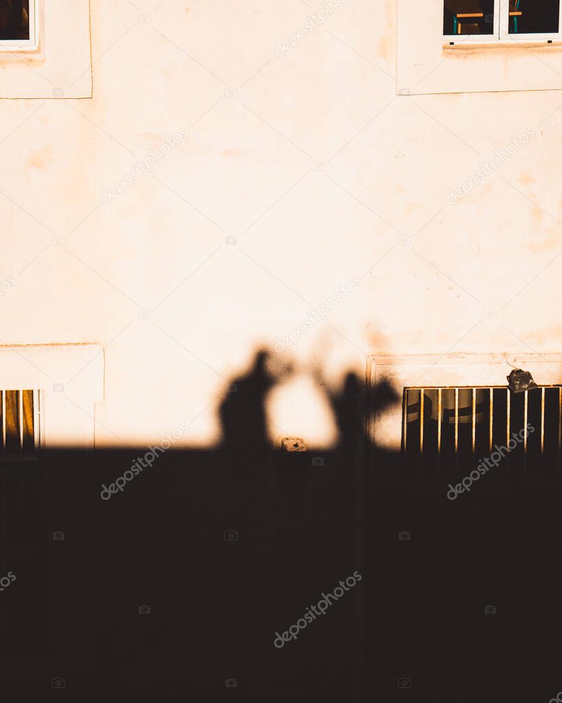Outlines, sihouettes as shadows are cast on a stone house of two people standing together. One person taking the photo while the other one holds their hands in the air