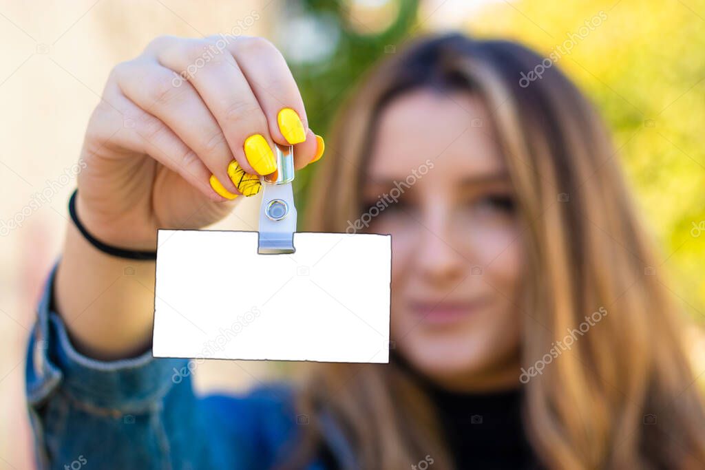Attractive brunette blurred in the background holding a blank nameplate card that can be filled out with information. Standing outside in nature on a bright day, fresh yellow beautiful manicured nails