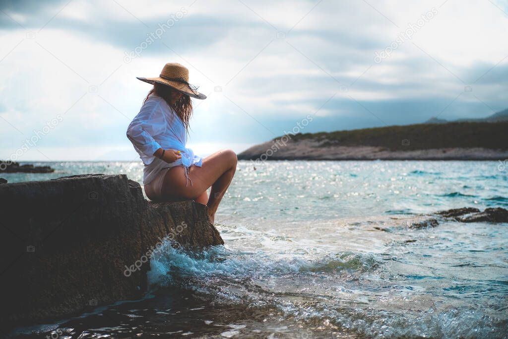 Side view of an amazing brunette sitting on a rock by the sea. Beautiful teal adriatic sea on the island of Vis, summer of 2021. Waves splashing on the shores